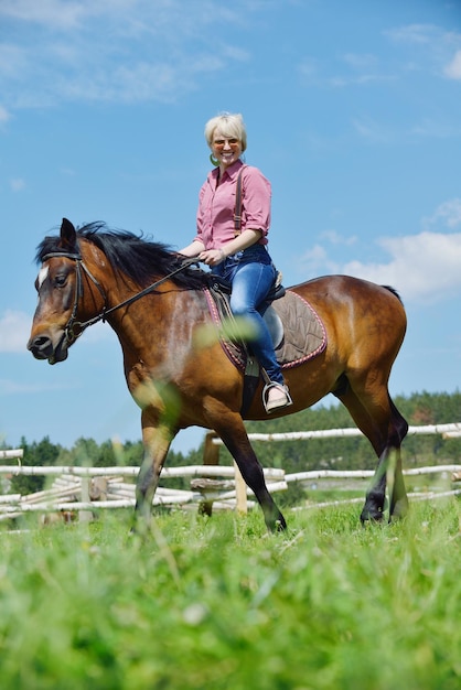 Glückliche Frau mit Sonnenbrille, die draußen auf einem Pferd auf dem Bauernhof sitzt, mit blauem Himmel im Hintergrund