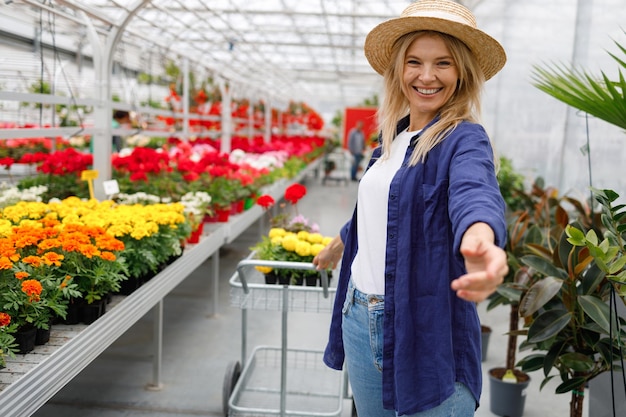 Glückliche Frau mit Strohhut geht in einem Gewächshaus mit einem Einkaufswagen für Blumen und andere Pflanzen spazieren