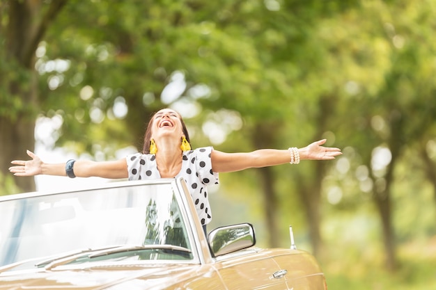 Glückliche Frau mit weit geöffneten Armen, die die Freiheit auf einem Cabrio-Auto-Roadtrip genießt.