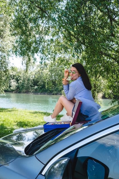 Glückliche Frau sitzt auf der Motorhaube und isst Pizza und schaut an einem sonnigen Sommertag auf den Fluss