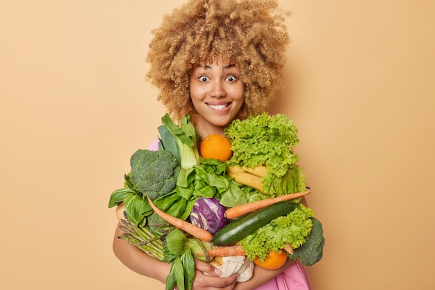 Glückliche Frau trägt frisches Bio-Gemüse und Obst und holt Lebensmittel für die Zubereitung von Salat oder anderen vegetarischen Gerichten, die über braunem Hintergrund isoliert sind Gesunde Ernährung und saisonales Erntekonzept