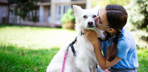 Glückliche Frau umarmt und küsst ihren weißen flauschigen Hund im Freien im Park auf dem Rasen Adoption gerettete Unterkunft Begleiter Haustier bester Freund