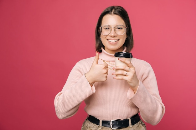 Glückliche Frau zeigt Daumen nach oben, während sie eine Kaffeetasse zum Mitnehmen hält Studioaufnahme über rosa Hintergrund