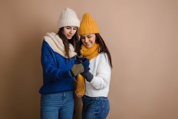 Glückliche Frauen der Schönheit in der Strickjacke und in Hüten, die Telefon auf braunem Hintergrund betrachten.