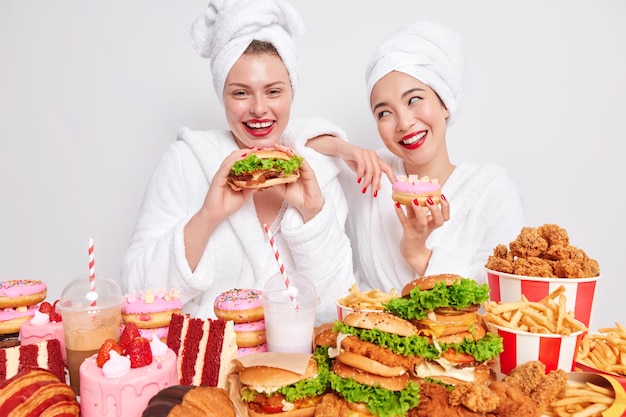 Glückliche Frauen genießen Cheat-Mahlzeit, essen leckere Burger-Kuchen und Donuts, die süchtig nach Fast Food sind