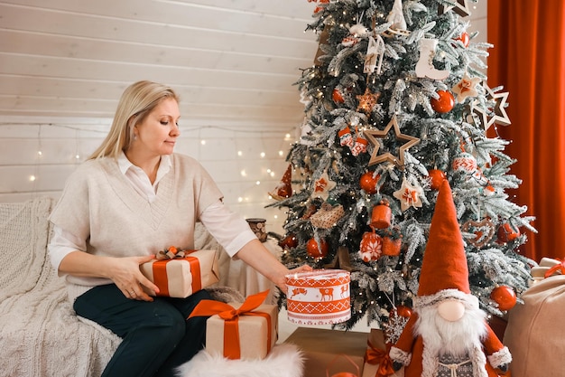 Glückliche Frauenmutter bereitet sich auf Weihnachten vor, Neujahr packt Geschenke zu Hause, während sie in der Nähe des Weihnachtsbaums sitzt. Winterurlaub, Dekorationskonzept