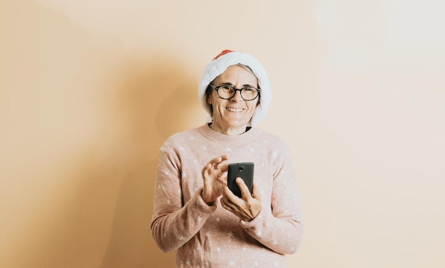 Glückliche fröhliche ältere alte weiße Haarfrau, die Weihnachtsmann-Hut trägt, lächelt, während sie intelligentes Telefon verwendet. steht vor einem farbigen Hintergrund. Weihnachtskleidung tragen. Platz kopieren. Platz für Werbung