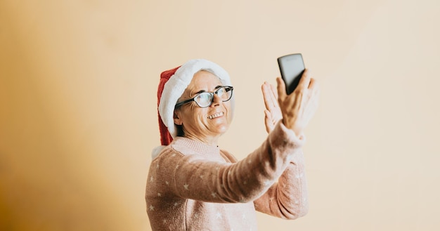 Glückliche fröhliche ältere alte weiße Haarfrau, die Weihnachtsmann-Hut trägt, lächelt, während sie intelligentes Telefon verwendet. steht vor einem farbigen Hintergrund. Weihnachtskleidung tragen. Platz kopieren. Platz für Werbung