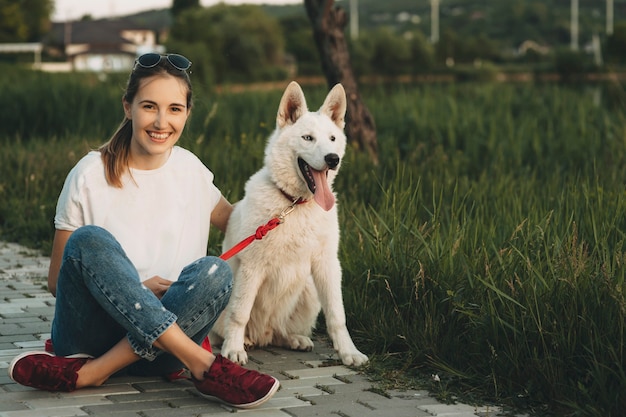 Glückliche fröhliche Frau mit gekreuzten Beinen, die auf Pflaster sitzen, großen weißen Hund umarmend und Kamera betrachten