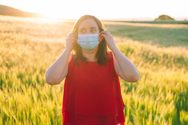 Glückliche fröhliche junge Frau, die medizinische Gesichtsmaske entfernt, die während des Sonnenuntergangs draußen auf der Wiese steht