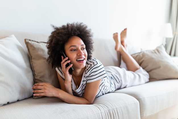 Glückliche fröhliche junge Frau, die zu Hause am Telefon spricht, lächelndes jugendlich Mädchen, das Anrufanruf durch Handy macht, das auf Sofa sitzt