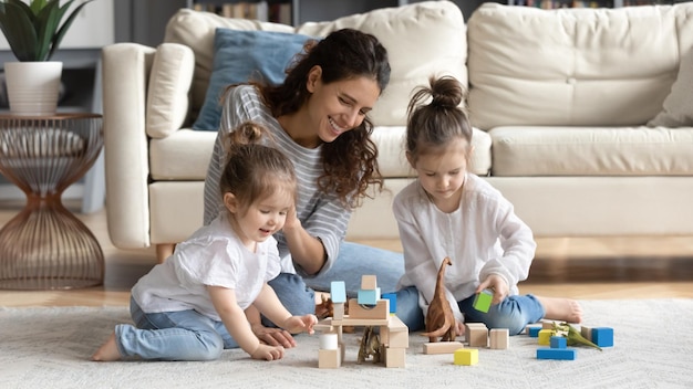 Glückliche fürsorgliche junge weiße Mutter haben Spaß spielen mit kleinen Tochter zu Hause im Wohnzimmer Lächeln liebevolle Mutter fühlen sich spielerisch engagiert in Spielaktivität mit Blöcken Ziegelsteine mit kleinen Mädchen Kinder