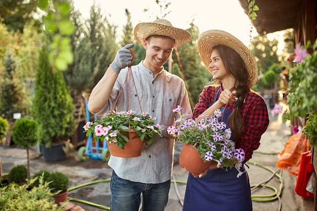 Glückliche Gärtner und Gärtner in Strohhüten halten an einem sonnigen Tag Töpfe mit Petunien auf dem Gartenweg. .