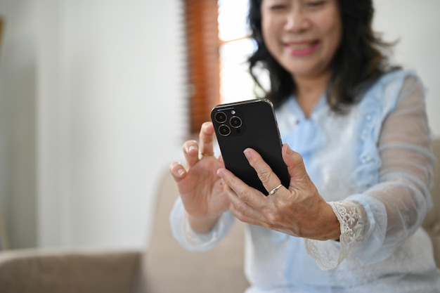 Glückliche gealterte asiatische Frau, die ihr Smartphone im selektiven Fokus des Wohnzimmers verwendet