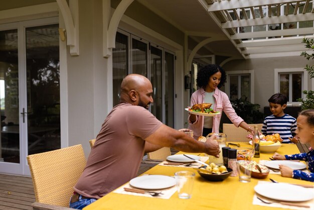 Glückliche gemischtrassige Eltern, Sohn und Tochter servieren Essen und sitzen zum Essen am Tisch auf der Terrasse. Sommer, Familie, Zweisamkeit, Essen und Lebensstil, unverändert.