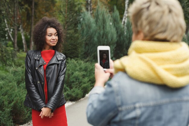 Glückliche gemischtrassige Freundinnen, die draußen Fotos auf dem Smartphone machen. Lächelnde Mädchen in lässigen warmen Outfits, die in der kalten Jahreszeit im Park spazieren gehen. Konzept für Technologie, Freundschaft und urbanes Stadtleben