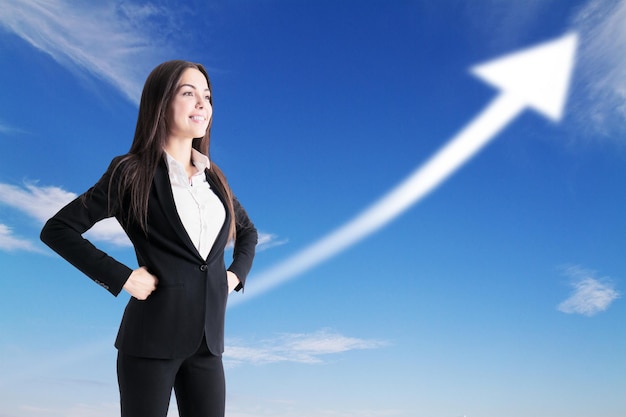 Glückliche Geschäftsfrau, die mit einem Pfeil nach oben im Hintergrund mit klarem Himmel steht Erfolgs- und Führungskonzept