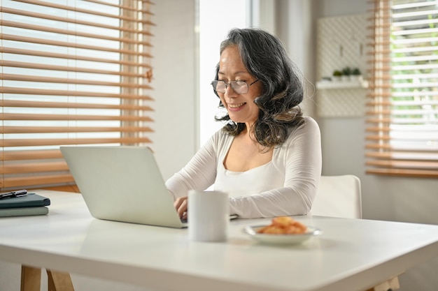 Glückliche Geschäftsfrau im asiatischen Alter der 60er Jahre, die mit ihrem Laptop von zu Hause aus arbeitet