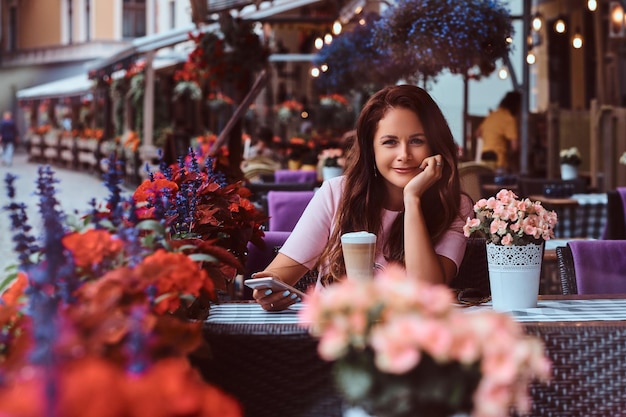 Glückliche Geschäftsfrau mittleren Alters mit langen braunen Haaren, die ein rosafarbenes Kleid trägt, hält Smartphone, während sie im Café im Freien sitzt.