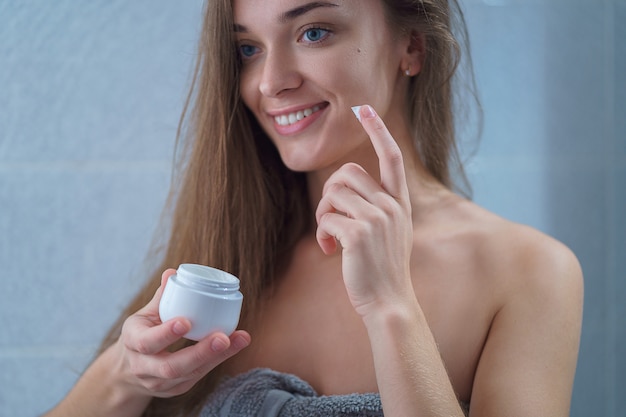 Glückliche gesunde attraktive brünette junge Frau im Handtuch tragen Feuchtigkeitscreme auf Gesicht im Badezimmer nach Dusche zu Hause auf