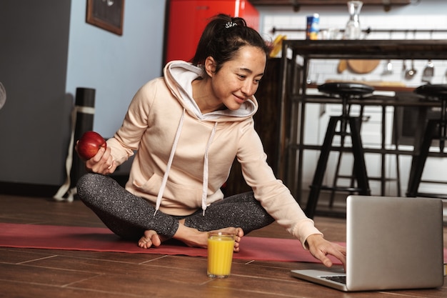 Glückliche gesunde Frau, die auf einer Fitnessmatte sitzt und Laptop nach dem Yoga-Training zu Hause verwendet