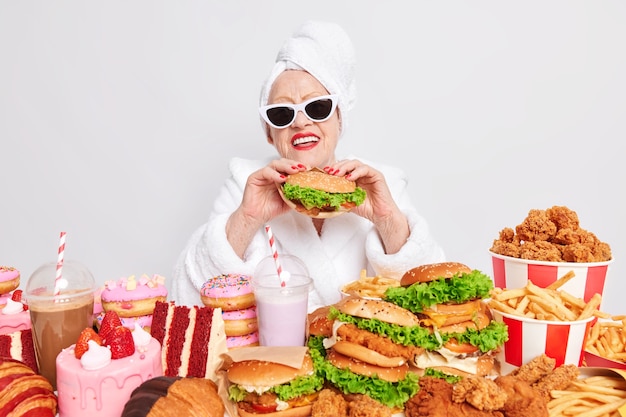 Glückliche Großmutter mit Sonnenbrille hält einen Hamburger umgeben von Fast Food