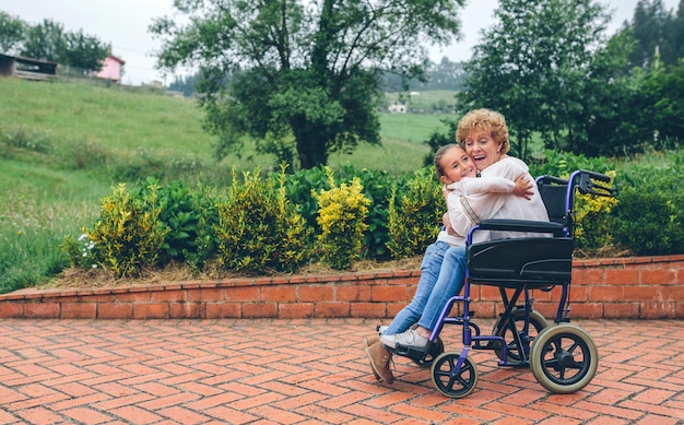 Foto glückliche großmutter umarmt schöne enkelin, während sie im rollstuhl im park sitzt