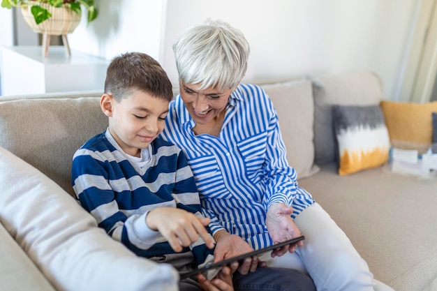 Glückliche Großmutter und Enkel, die zusammen Tablet verwenden, auf einem gemütlichen Sofa zu Hause sitzen, Tablet-Geräte-Apps durchsuchen, Oma mit Enkelkind spielen, auf den Bildschirm schauen, Spaß haben