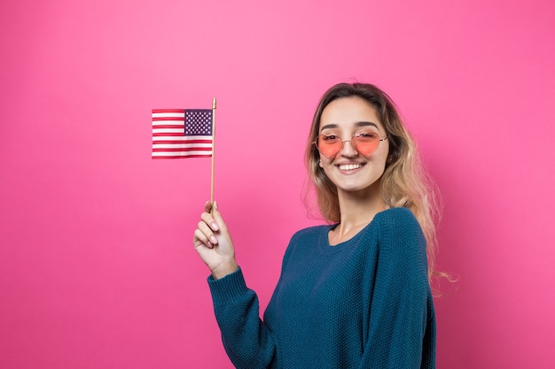 Glückliche herzförmige Brille der jungen Frau, die amerikanische Flagge gegen einen rosafarbenen Hintergrund des Studios hält