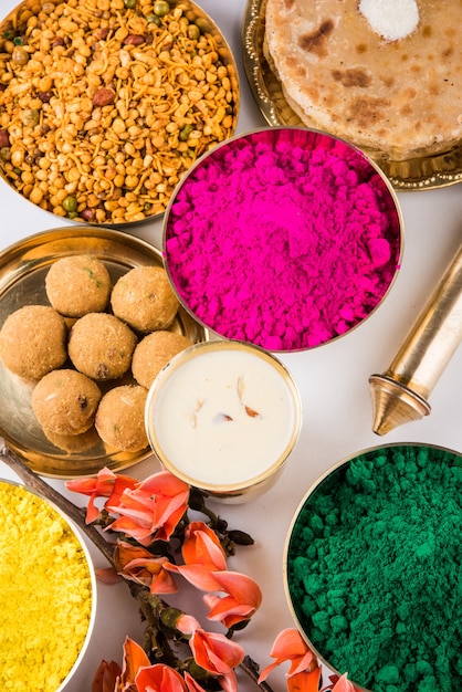 Glückliche Holi-Grußkarte, die indisches traditionelles süßes und salziges Essen, Blumen und Pulverfarben zeigt, die über weißem Hintergrund oder Ton angeordnet sind. Selektiver Fokus