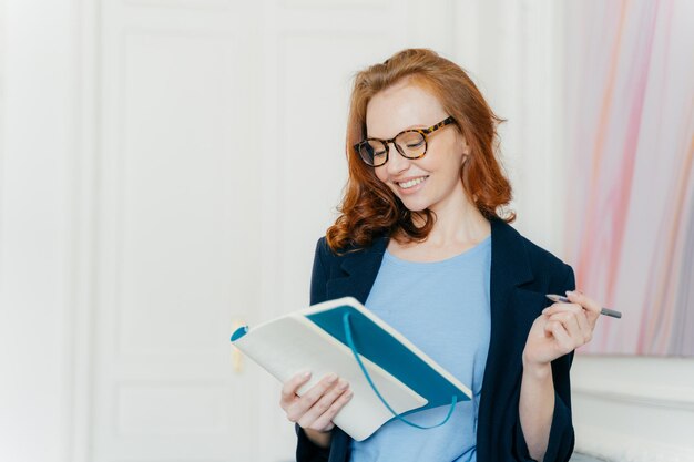 Glückliche Ingwer junge Frau plant Arbeitsplan schreibt in Notizbuch macht Notizen von nützlichen Informationen hält Stift hat ein Lächeln im Gesicht elegant gekleidet steht drinnen schreibt ihre Gedanken auf