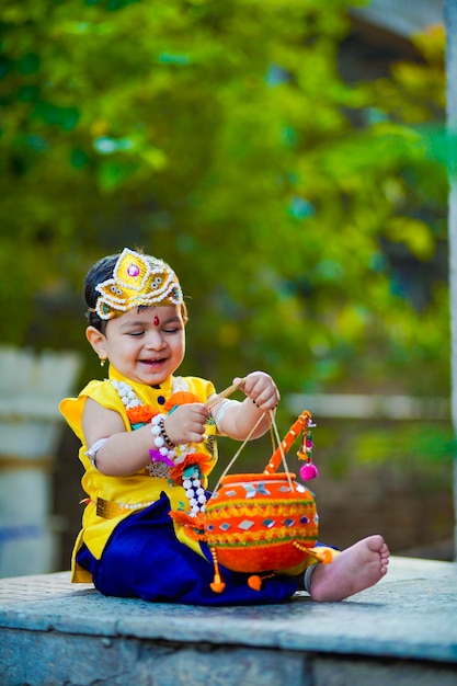 Glückliche Janmashtami-Grußkarte, die den kleinen indischen Jungen zeigt, der als Shri Krishna oder Kanha/Kanhaiya mit Dahi Handi-Bild und bunten Blumen aufwirft