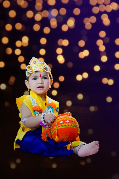 Glückliche Janmashtami-Grußkarte, die den kleinen indischen Jungen zeigt, der als Shri Krishna oder Kanha/Kanhaiya mit Dahi Handi-Bild und bunten Blumen aufwirft