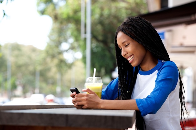 Glückliche junge afrikanische Frau, die Café am im Freien unter Verwendung des Mobiltelefons sitzt