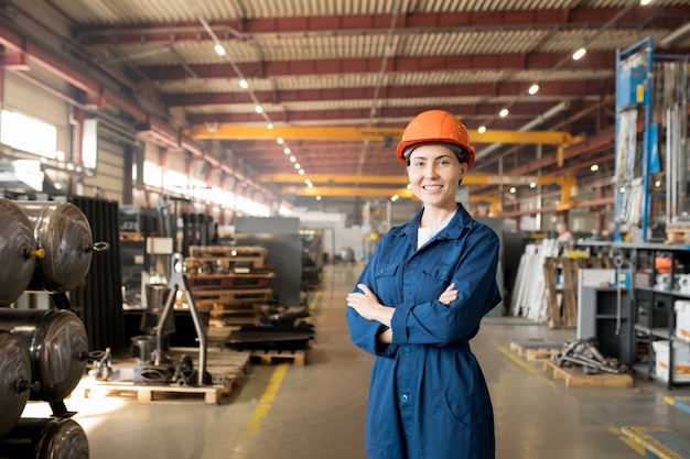 Glückliche junge Arbeiterin der großen modernen Fabrik in blauer Uniform und Schutzhelm, die ihre Arme auf Brust kreuzen