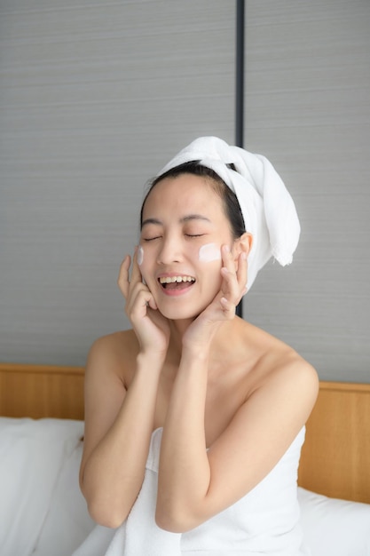 Glückliche junge asiatische Frau, die Gesichtslotionen aufträgt, während sie ein Handtuch trägt und ihr Gesicht berührt Tägliches Make-up und Hautpflege