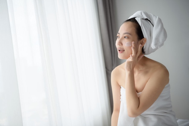 Glückliche junge asiatische Frau, die Gesichtslotionen aufträgt, während sie ein Handtuch trägt und ihr Gesicht berührt Tägliches Make-up und Hautpflege