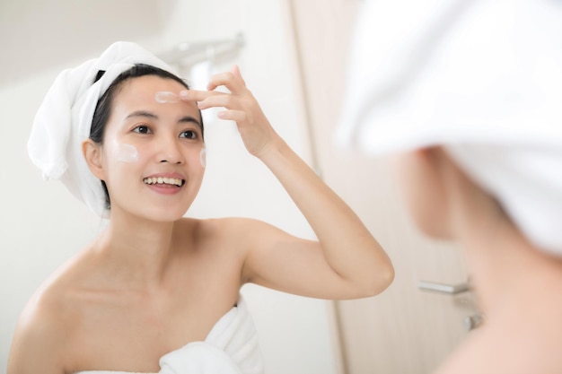 Glückliche junge asiatische Frau, die Gesichtslotionen aufträgt, während sie ein Handtuch trägt und ihr Gesicht im Badezimmer berührt