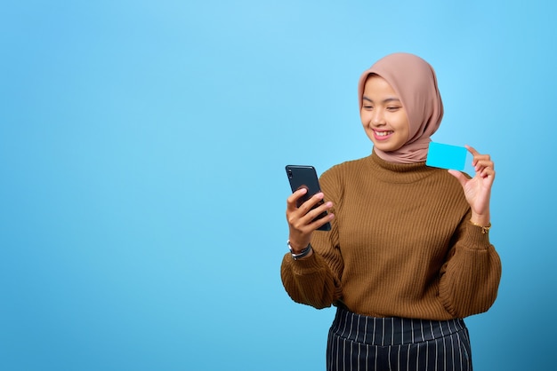 Glückliche junge asiatische Frau, die Kreditkarte und Handy zur Hand auf blauem Hintergrund zeigt