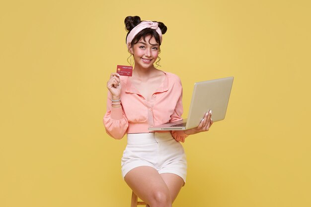 Glückliche junge asiatische Frau, die Kreditkarte zeigt und Computer-Online-Shopping auf gelbem Hintergrund verwendet