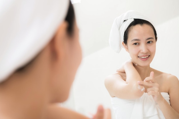 Glückliche junge asiatische Frau, die Lotionen auf ihrem Körper im Badezimmer anwendet