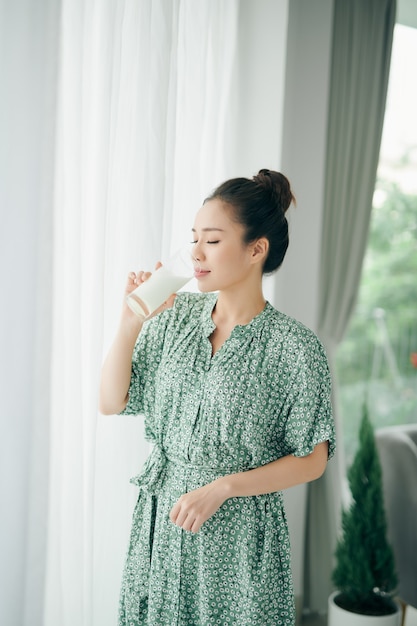 Glückliche junge asiatische Frau, die Milch vom Glas im Wohnzimmer zu Hause trinkt