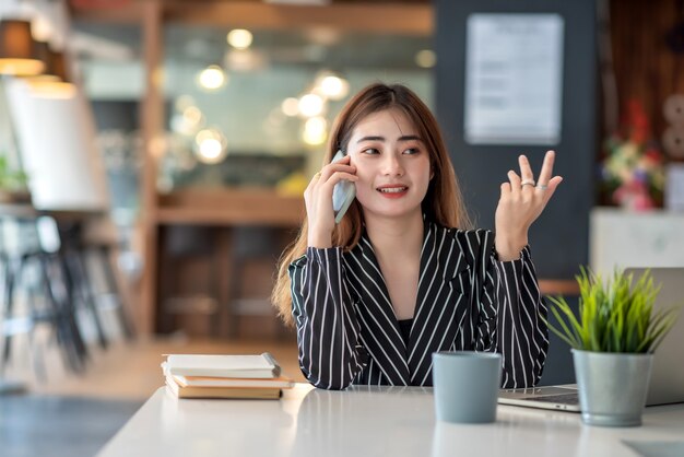 Glückliche junge asiatische Geschäftsfrau, die am Laptop arbeitet und auf Smartphone spricht