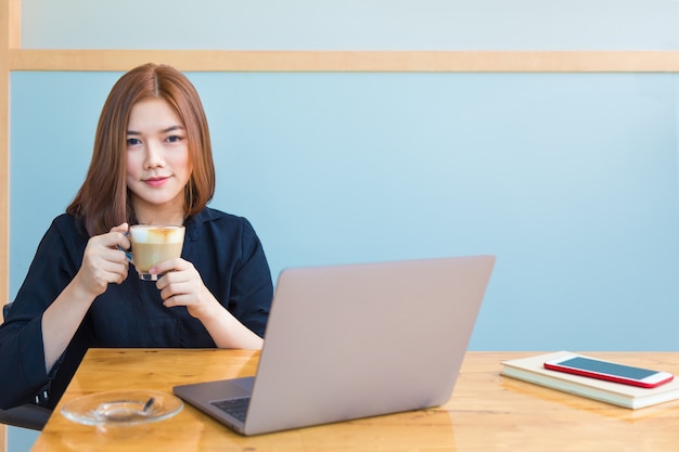 Glückliche junge asiatische Geschäftsfrau, die ihren heißen Kaffee beim Arbeiten genießt