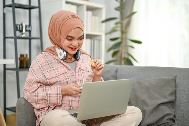 Glückliche junge asiatische muslimische Frau, die Kekse isst und Filme auf ihrem Laptop im Wohnzimmer anschaut