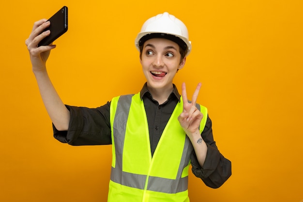 Glückliche junge Baumeisterin in Bauweste und Schutzhelm, die fröhlich lächelt und Selfie mit dem Smartphone macht, das ein V-Zeichen auf Orange zeigt