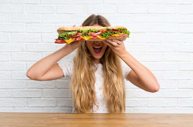 Glückliche junge blonde Frau, die ein großes Sandwich anhält