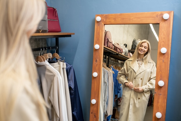 Glückliche junge blonde Frau, die neuen eleganten beigen Mantel vor dem Spiegel in der Umkleidekabine anprobiert, während sie neue Kleidung für den Frühling wählt