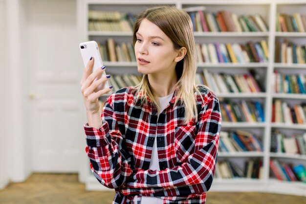 Glückliche junge blonde Studentin, die lässiges kariertes Hemd trägt, in der modernen Bibliothek steht und Textnachricht liest oder mit ihrem Handy studiert