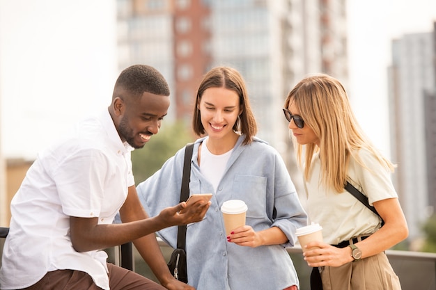 Glückliche junge blonde und brünette Frauen in Freizeitkleidung, die Getränke trinken, während sie die Anzeige des Smartphones in der Hand eines gutaussehenden afrikanischen Mannes betrachten